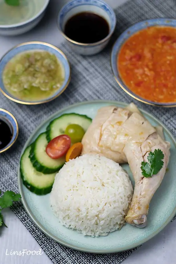 Hainanese Chicken Rice on a plate with chicken leg, cucumbers, tomatoes and dips of chilli sauce and ginger and spring onion and soy sauce