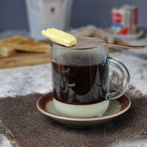 butter coffee (kopi gu you) in a glass mug, showing a layer of black coffee over a layer of condensed milk and a cube of butter resting on a spoon