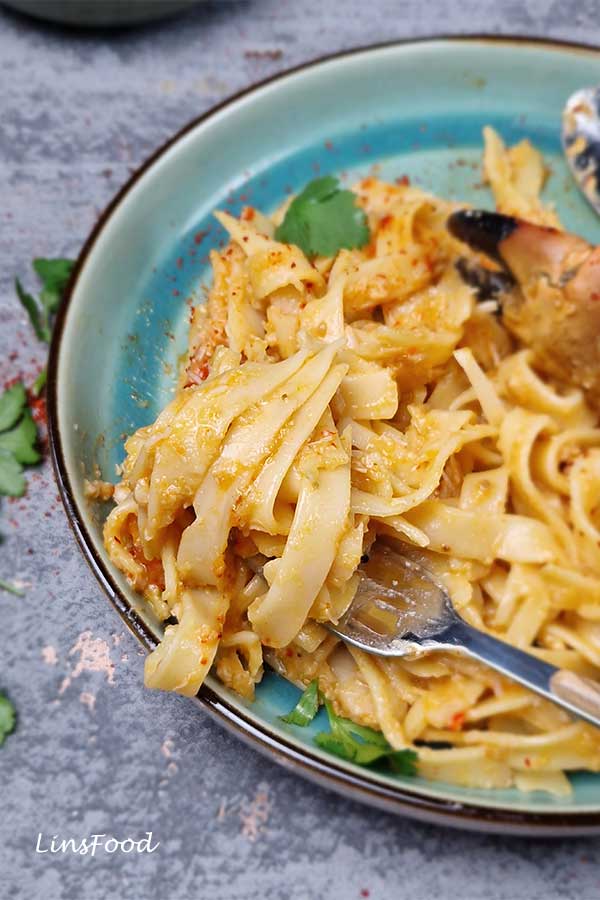 Close-up of chilli crab pasta twirled on a fork over a blue-green plate, thick sweet-spicy Singapore chilli crab sauce and crab meat.
