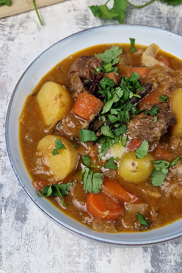 Close-up of hearty Eurasian beef stew in a light blue bowl, finished with butter and coriander leaves.