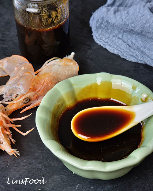 Dark homemade petis udang on a white spoon in a green bowl with prawn shells behind