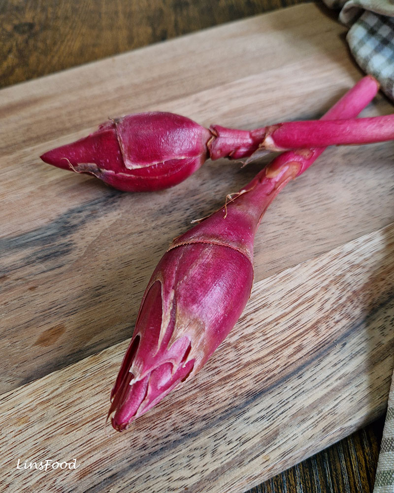 Two vivid pink bunga kantan flower buds on a wooden board, one positioned close to the camera and the other behind it.