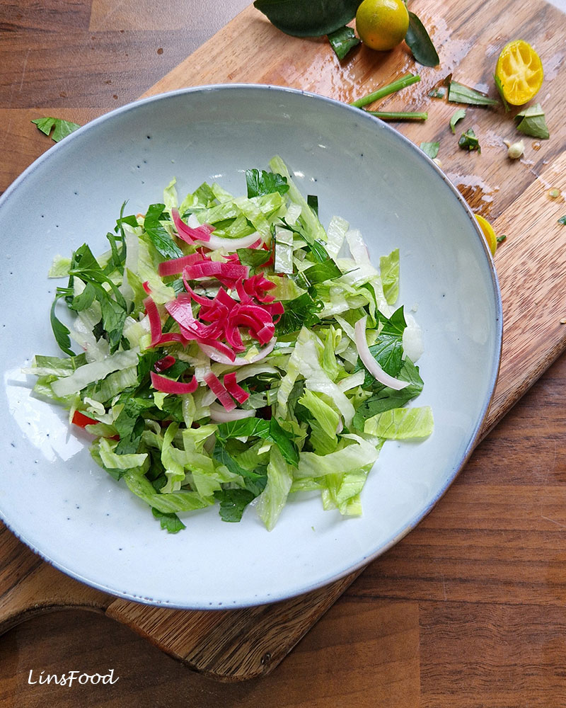Green herb and lettuce salad topped with thin strips of bright pink bunga kantan, with calamansi on the chopping board behind.