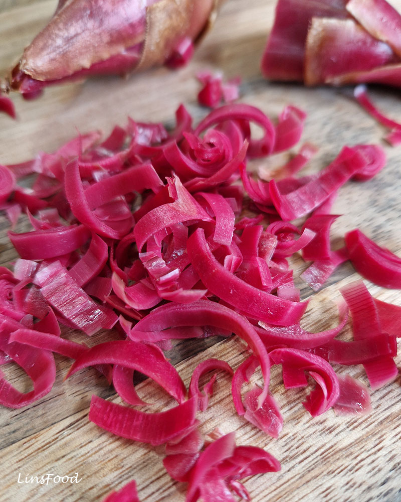 Close-up of thinly sliced bright pink bunga kantan scattered on a wooden chopping board.