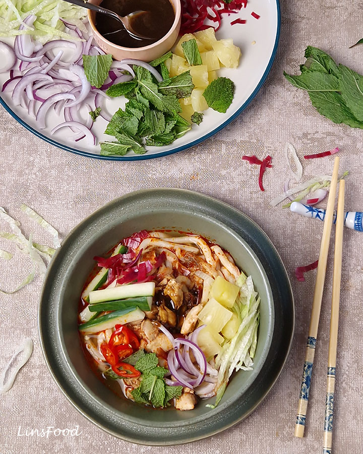 Bowl of Penang asam laksa with chopsticks and fresh toppings alongside