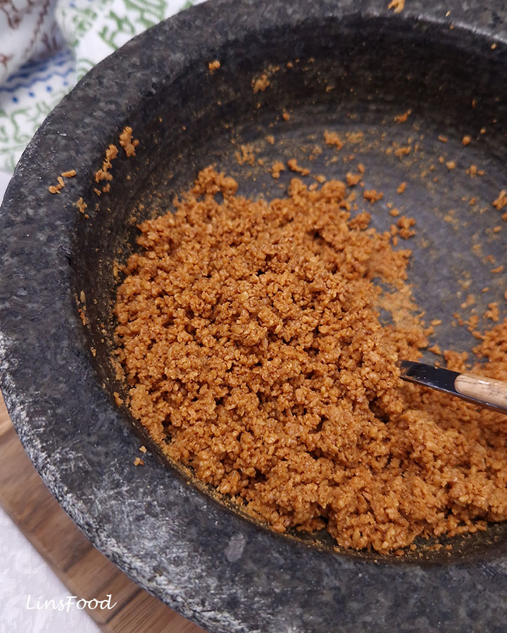 Golden-brown kerisik gathered into crumbly clumps in a dark stone mortar, with a spoon entering from the right.