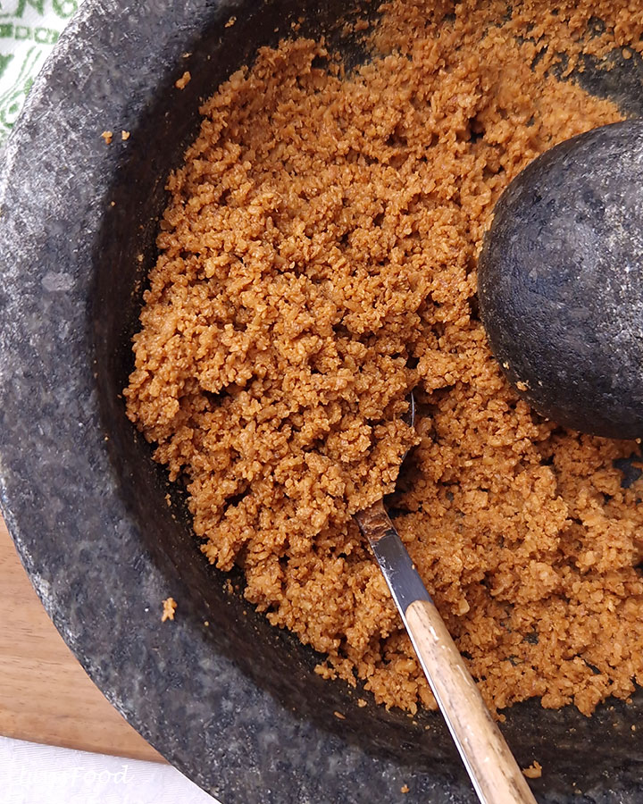 Close-up of a spoonful of golden-brown kerisik beside a stone pestle, showing fine coconut crumbs clumped together.